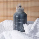 A dark gray bottle of FIRSTHAND body wash with a clear cap, nestled in a white textured towel with a warm, blurred wooden background.