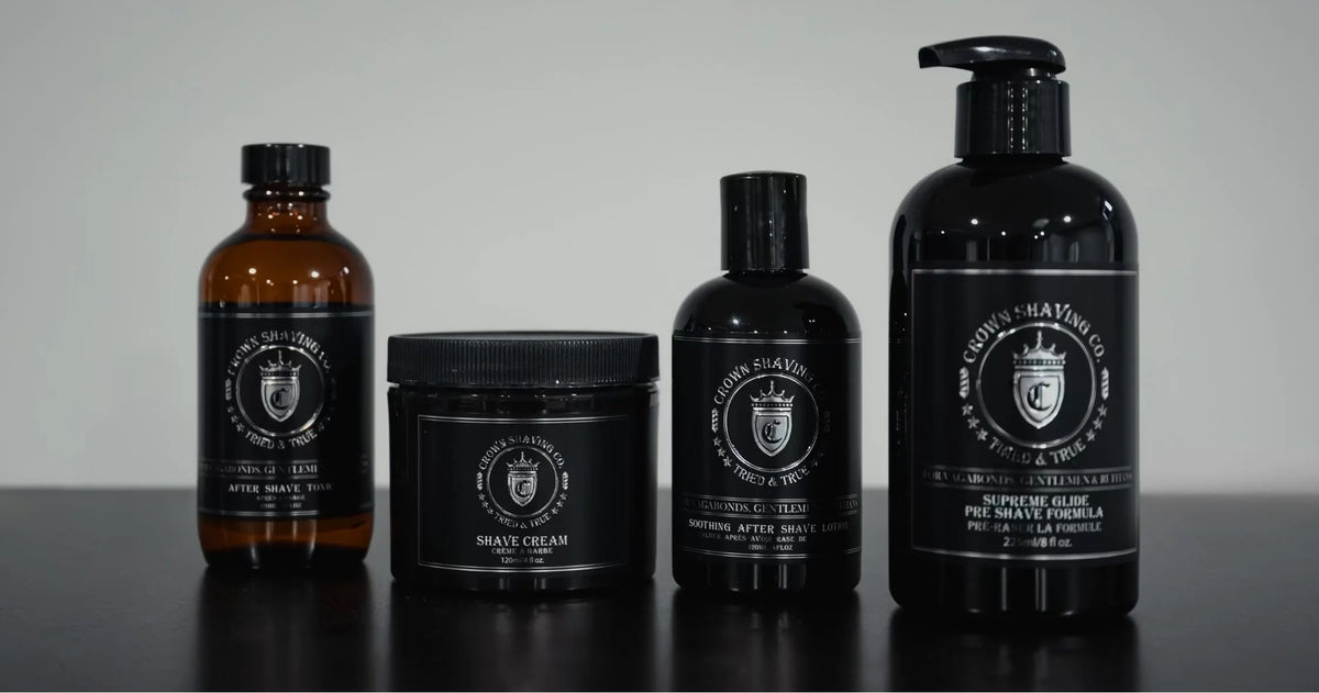 Four grooming products from Global Grooming are lined up on a dark surface against a light gray background. The set includes an amber glass bottle, a black jar of shave cream, a black bottle of shaving soap, and a large black pump bottle of shaving lotion.