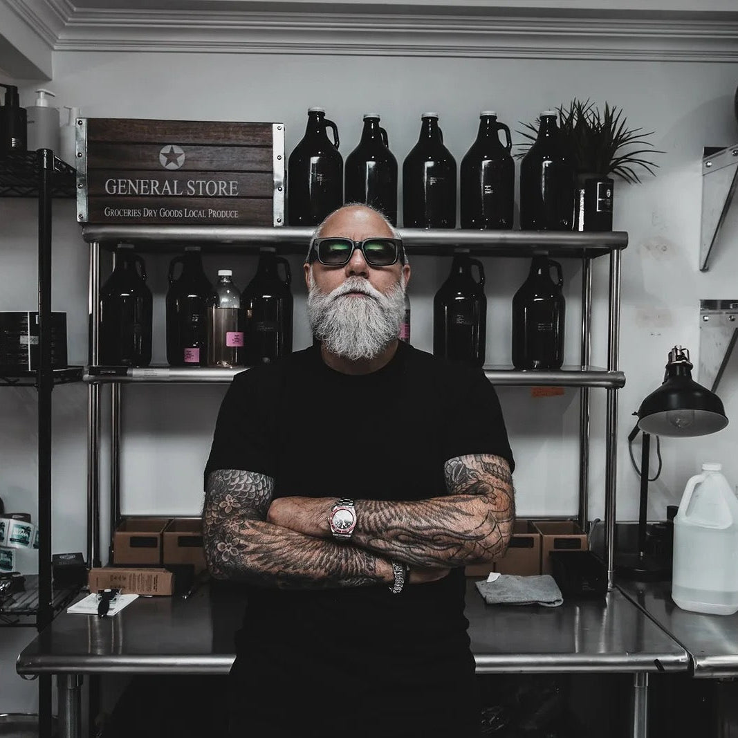 A man with a gray beard, tattoos, and sunglasses stands with arms crossed in a workshop or retail space, with shelves of bottles and products behind him.