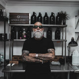 A man with a gray beard, tattoos, and sunglasses stands with arms crossed in a workshop or retail space, with shelves of bottles and products behind him.